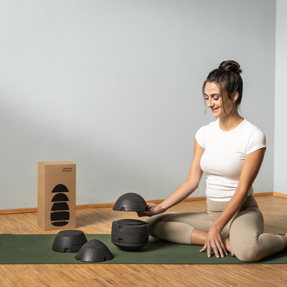 Eine Frau in Trainingskleidung sitzt auf einer Yogamatte im Haus, lächelt und hält einen schwarzen Balance-Dome. Neben ihr auf dem Holzboden stehen mehrere Balance-Kuppeln und ein Karton.