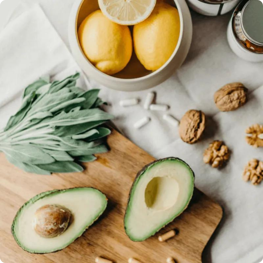 Eine halbierte Avocado, eine Schale mit zwei Zitronen, Walnüsse, grüne Blattkräuter und verschiedene Kapseln und Pillen sind auf einem hölzernen Schneidebrett und einer weißen Fläche in der Nähe angeordnet.