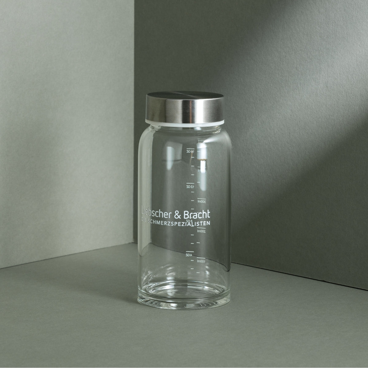 Eine Liebscher & Bracht Glasflasche mit Metalldeckel sitzt auf einer grauen Fläche, mit weißen Maßen und "Liebscher & Bracht SCHMERZSPEZIALISTEN" auf der Vorderseite - perfekt für Mixen von Pulver oder Nahrungsergänzungsmittel.