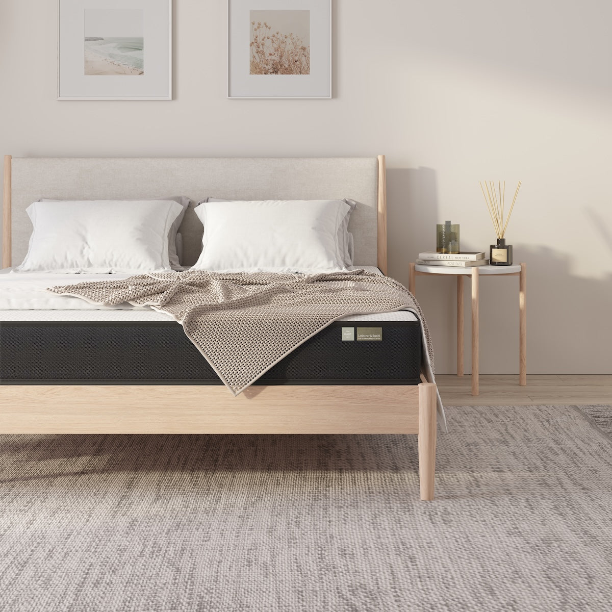 Ein ordentlich gemachtes Bett mit einer beigen Strukturdecke und weißen Kissen auf der Liebscher & Bracht Matratze auf einem hellen Holzrahmen sorgt für optimalen Komfort. Ein Nachttisch, Deko-Akzente, ein hellgrauer Teppich und zwei gerahmte Bilder vervollständigen die Szene.