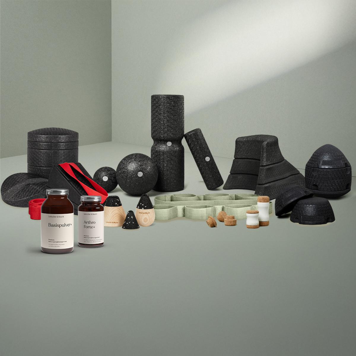 Eine Vielzahl von schwarzen Schaumstoffrollen, Massagebällen, Balancepads, zwei Nahrungsergänzungsflaschen, roten Widerstandsbändern und kleinen Korkgläsern sind auf einer hellgrauen Fläche vor einem gedämpften grauen Hintergrund angeordnet.