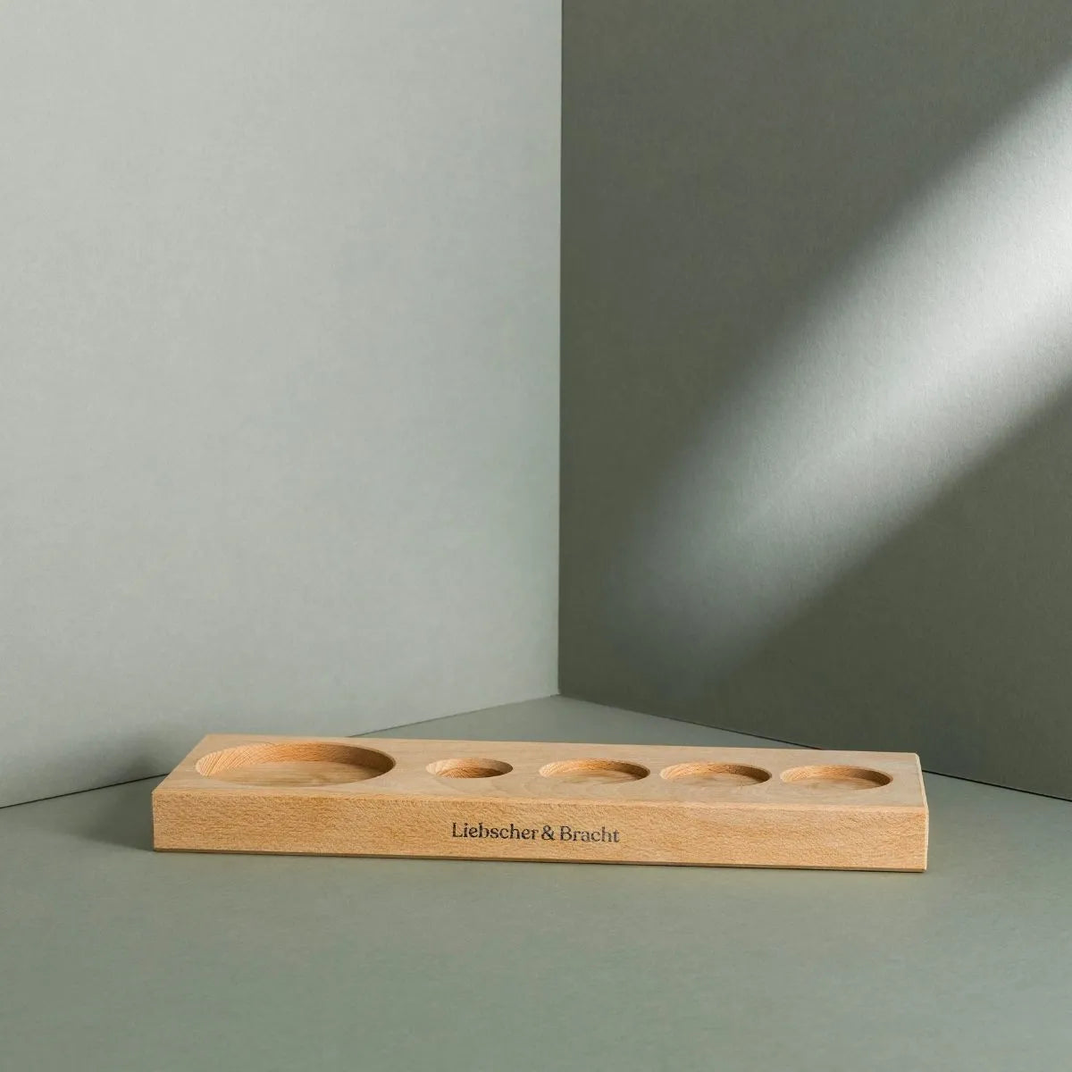 Ein Liebscher & Bracht Aufbewahrungsbrett mit fünf unterschiedlich großen kreisrunden Vertiefungen hält ein Drücker-Set auf einer grauen Fläche und Wand, der Markenname ist auf der Vorderseite aufgedruckt, während weiches Licht sanfte Schatten wirft.