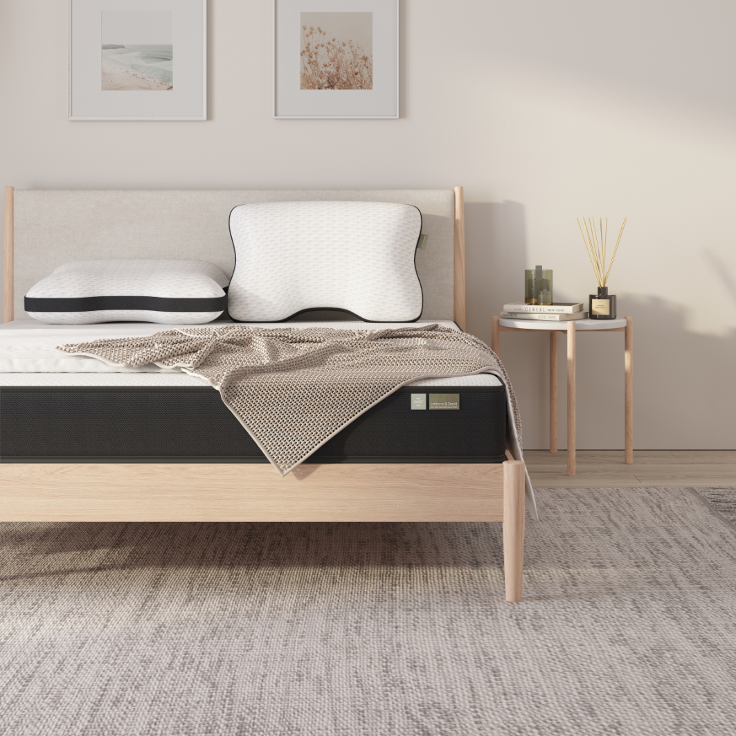 Minimalistisches Schlafzimmer mit einem Holzbett, zwei Kissen - darunter das Liebscher & Bracht Kissen zur optimalen Unterstützung des Rückens -, einer strukturierten beigen Decke, einem hellgrauen Teppich, gerahmter Wandkunst und einem kleinen Nachttisch mit Dekoration und Schilfrohrdiffusoren.