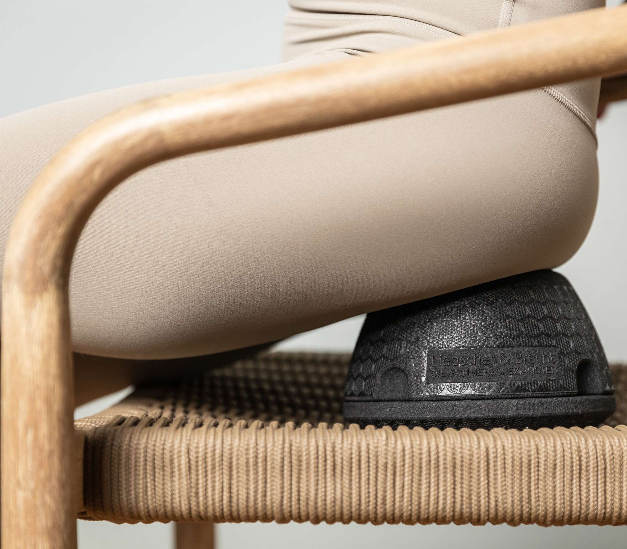 Eine Person in beiger Kleidung sitzt auf einem Holzstuhl mit geflochtener Sitzfläche und benutzt ein schwarzes, kuppelförmiges Kissen, das zur ergonomischen Unterstützung unter ihr liegt.
