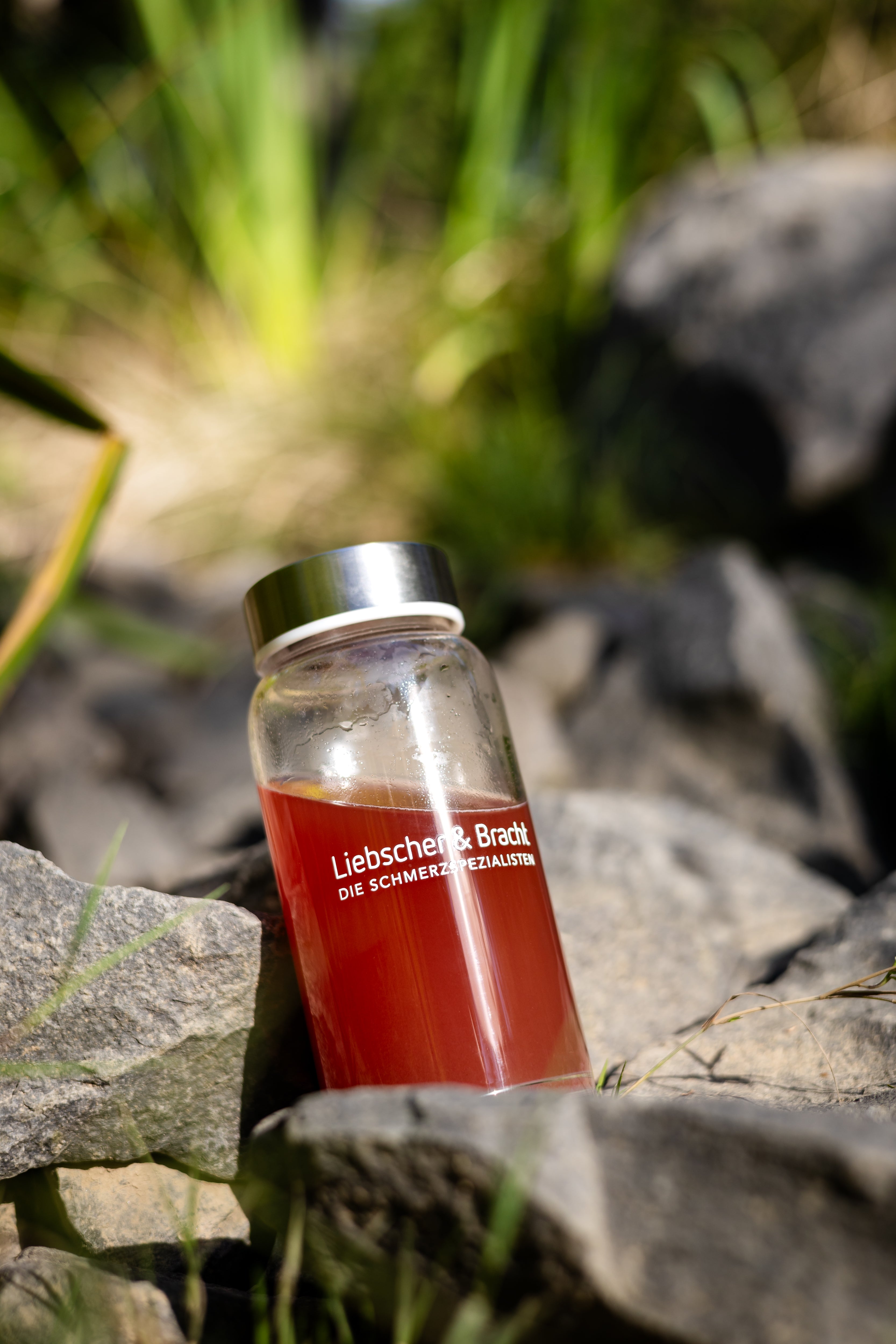 Eine Glasflasche mit Metalldeckel, die mit einer roten Flüssigkeit gefüllt ist und die Aufschrift Liebscher & Bracht DIE SCHMERZSPEZIALISTEN trägt, steht auf einem Felsen im Freien, mit grünem Gras im unscharfen Hintergrund.