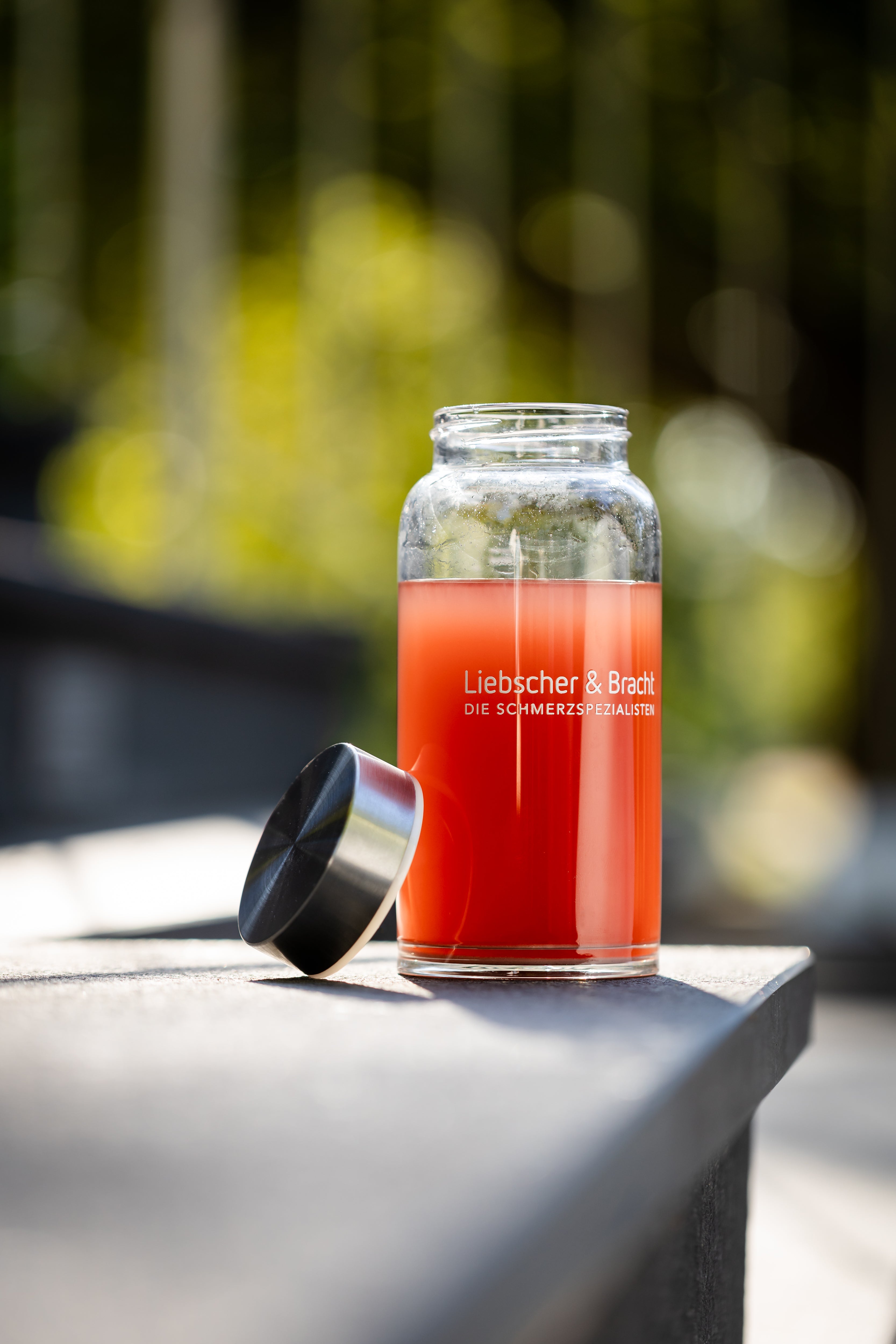 Ein mit roter Flüssigkeit gefülltes Glasgefäß mit der Aufschrift Liebscher & Bracht DIE SCHMERZSPEZIALISTEN steht auf einer sonnenbeschienenen Fläche im Freien, der Deckel liegt daneben; im Hintergrund ist verschwommenes Grün zu erkennen.