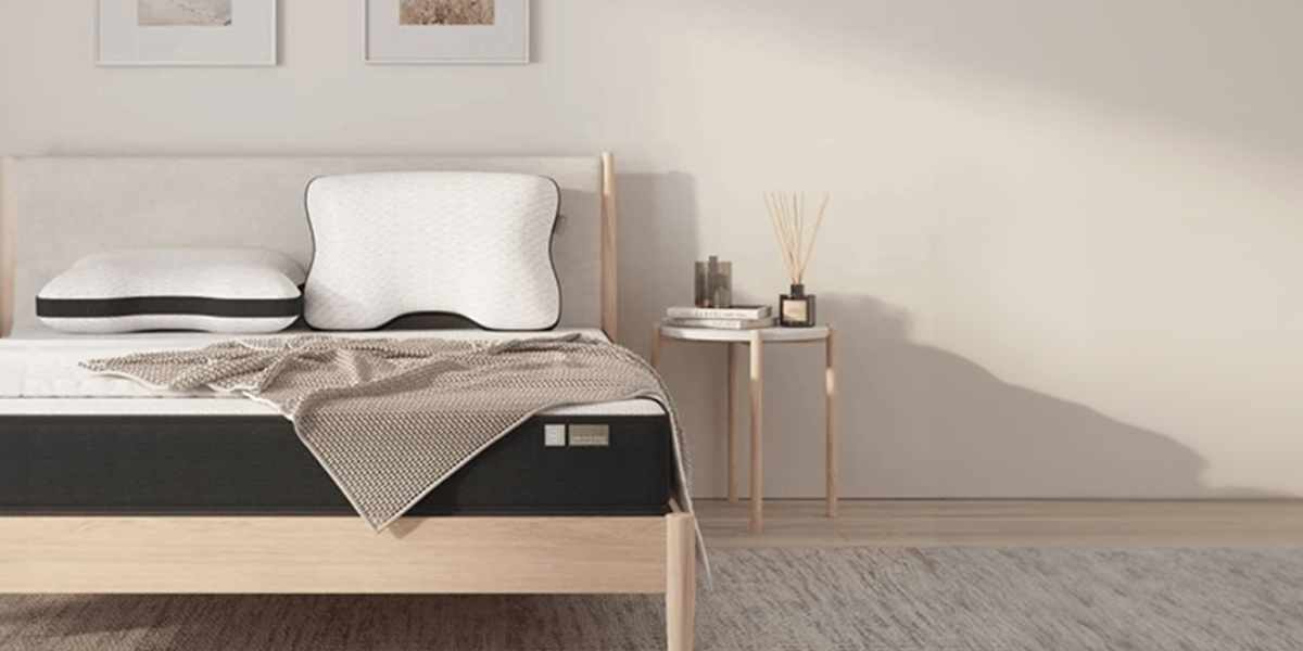 Ein ordentlich gemachtes Bett mit zwei ergonomischen Kissen, einer beigen Decke und einem hellen Holzrahmen steht neben einem kleinen runden Beistelltisch mit Dekorationsartikeln in einem hellen, modernen Schlafzimmer.