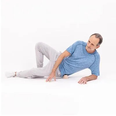 Ein Mann in blauem Hemd und grauer Hose liegt auf der Seite auf dem Boden, stützt sich auf einen Ellbogen und hält einen Massageball unter seine Hüfte, um eine Selbstmassageübung durchzuführen.