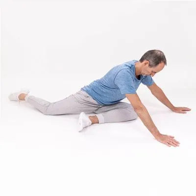 Ein Mann in blauem T-Shirt und grauer Hose dehnt sich im Sitzen auf dem Boden, wobei er ein Bein nach hinten beugt und das andere nach vorne streckt und beide Arme nach vorne streckt.