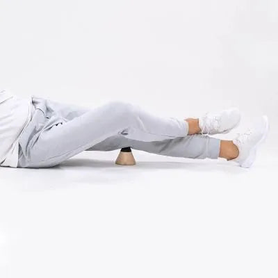 Eine Person in hellgrauer Hose und weißen Turnschuhen liegt auf dem Rücken und hält sich einen hölzernen Massageroller unter den Oberschenkel, vor einem schlichten weißen Hintergrund.