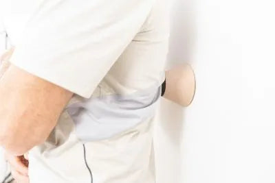 Eine Person in einem hellgrauen Hemd, die seitlich an einer weißen Wand steht und einen rosafarbenen konischen Gegenstand zwischen sich und die Wand presst.
