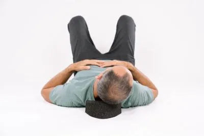 Ein Mann liegt auf dem Rücken mit angewinkelten Knien, die Hände ruhen auf dem Bauch, und sein Kopf wird von einer schwarzen Schaumstoff-Nackenstütze gestützt, auf einem schlichten weißen Hintergrund.