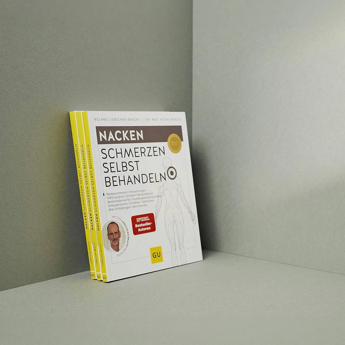 Drei Exemplare des deutschen Buches "Nackenschmerzen selbst behandeln" von Liebscher & Bracht, in dem es um ein Übungsprogramm für Nackenschmerzen geht, sind aufrecht an einer grauen Wand und auf dem Boden gestapelt. Der Umschlag enthält gelbe Akzente und ein Autorenfoto.