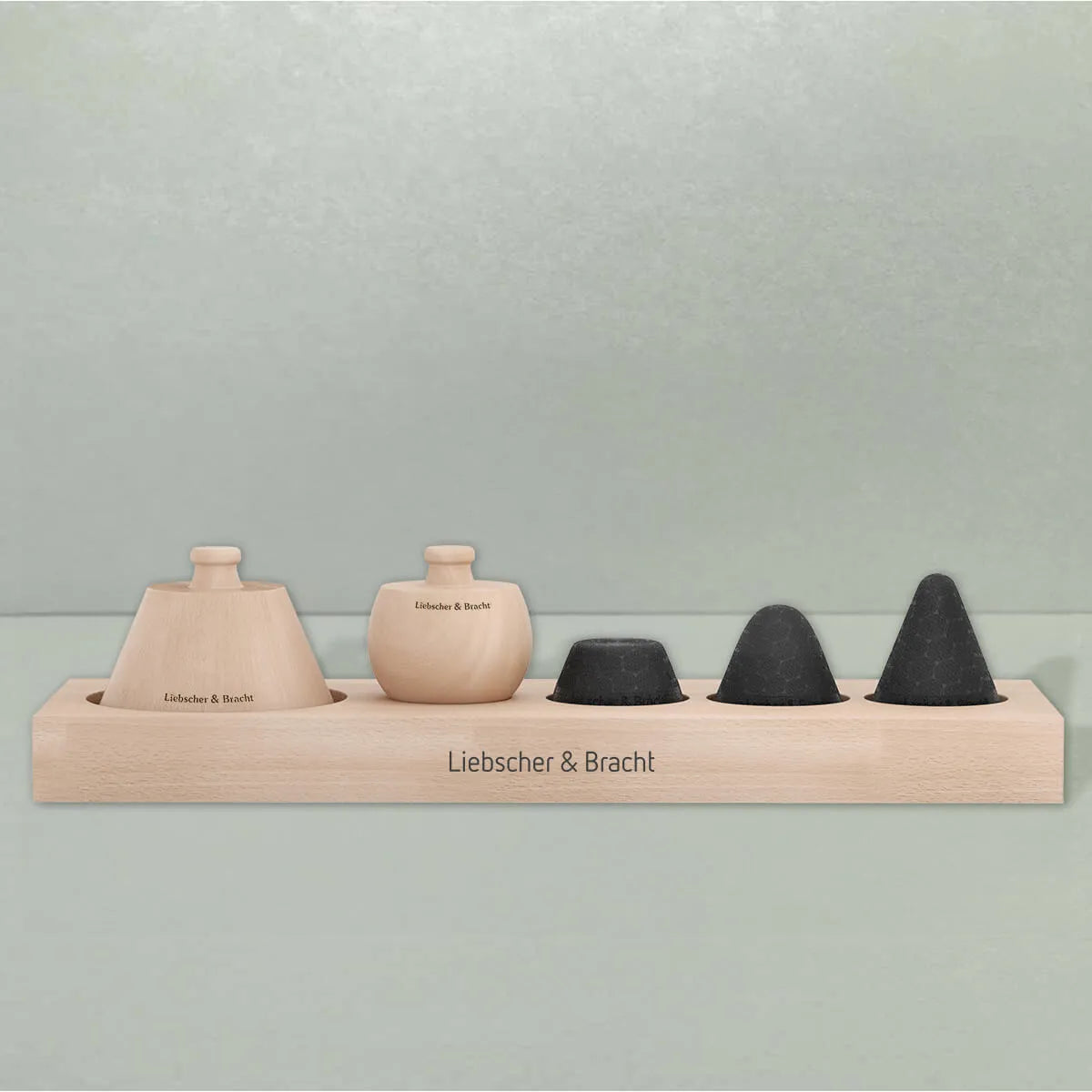 Das Aufbewahrungsbrett von Liebscher & Bracht fasst zwei unterschiedlich geformte Massagewerkzeuge aus hellem Holz und zwei schwarze, kegelförmige Massagesteine in einer Reihe, auf deren Holzsockel der Markenname aufgedruckt ist.