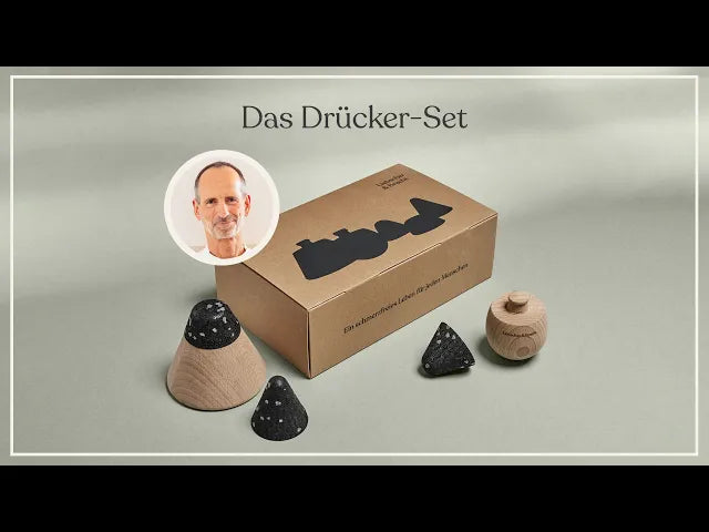 Eine Pappschachtel mit der Aufschrift Das Drücker-Set ist ausgestellt, vor der drei kegelförmige Objekte aus Holz und Stein stehen. In der oberen linken Ecke ist das Foto eines lächelnden Mannes eingefügt.