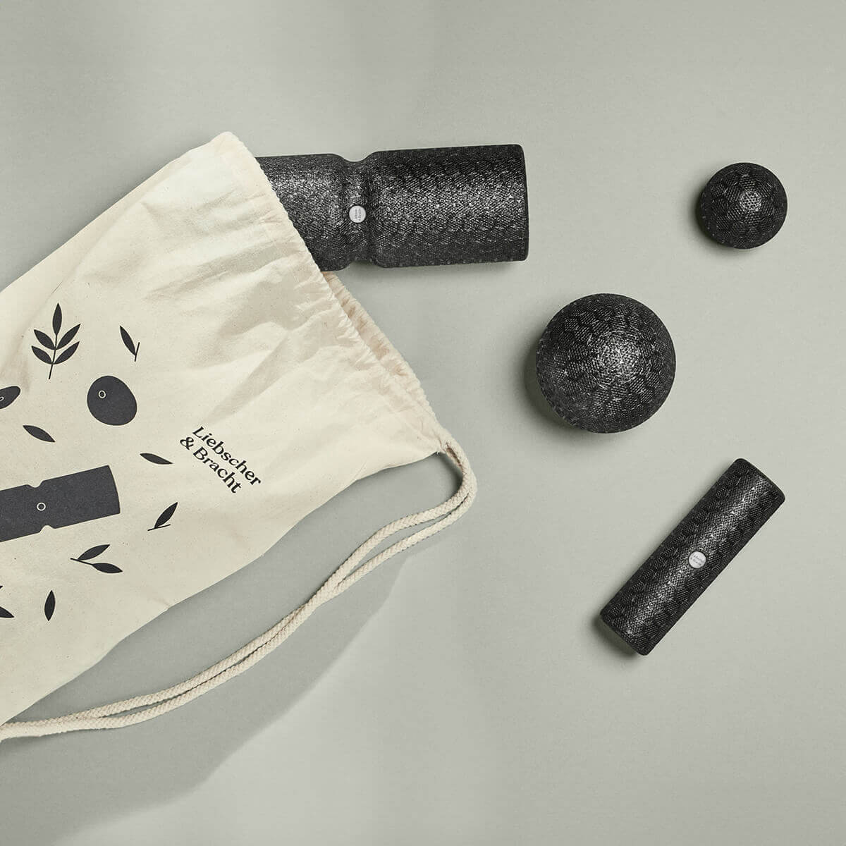 Ein beiger Kordelzugbeutel mit schwarzem, minimalistischem Design aus dem Unsere Empfehlung | Faszien-Set + Übungs-Schlaufe von Liebscher & Bracht liegt auf einer grauen Fläche, die teilweise geöffnet ist und den Blick auf strukturierte Faszienrollen und Massagebälle für effektive Dehnübungen freigibt.