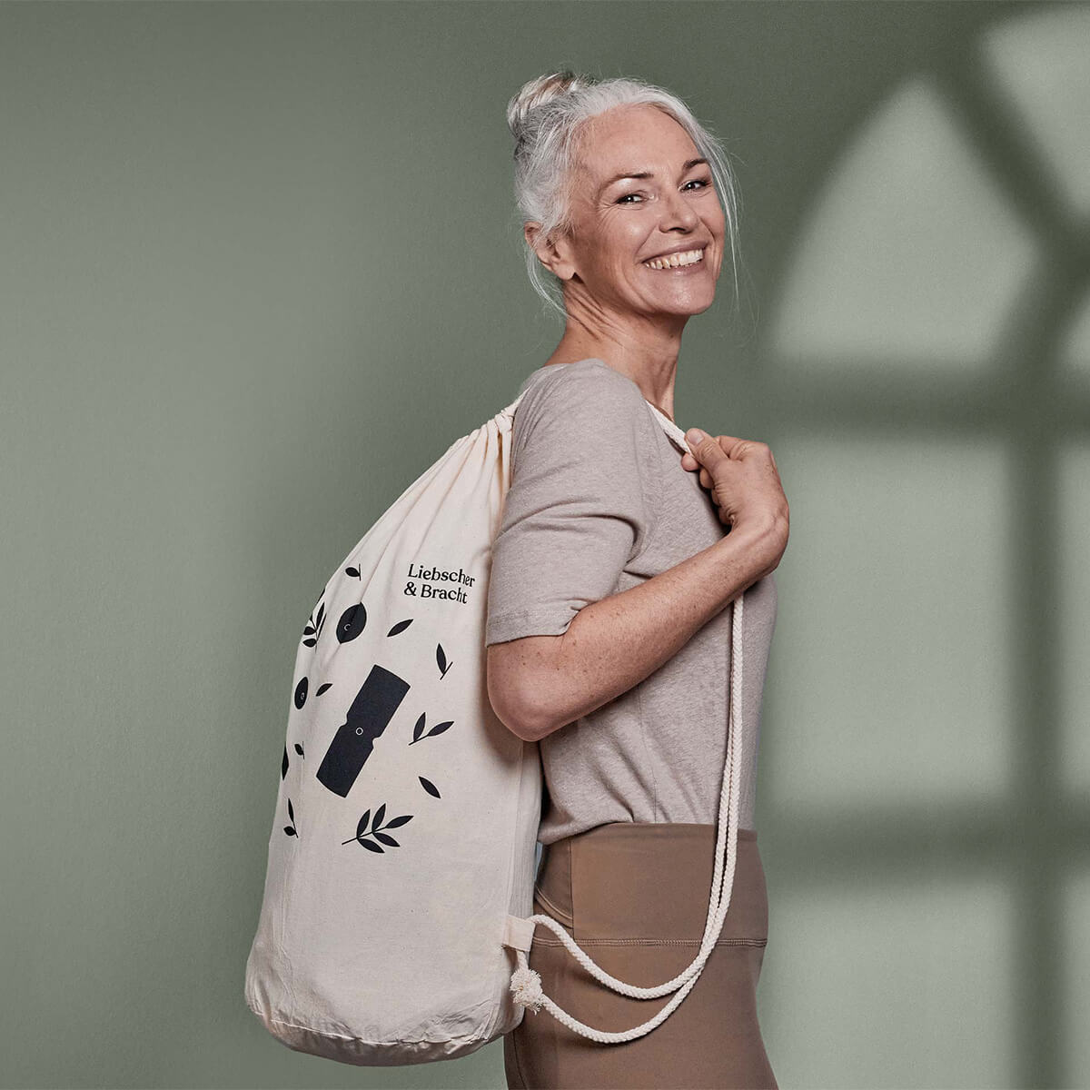 Eine lächelnde ältere Frau mit grauem Haar in einem Pferdeschwanz, die ein beiges Hemd und eine braune Hose trägt, steht seitlich und hält eine cremefarbene Kordeltasche mit schwarzen Mustern über ihre Schulter vor einem grünen Hintergrund.