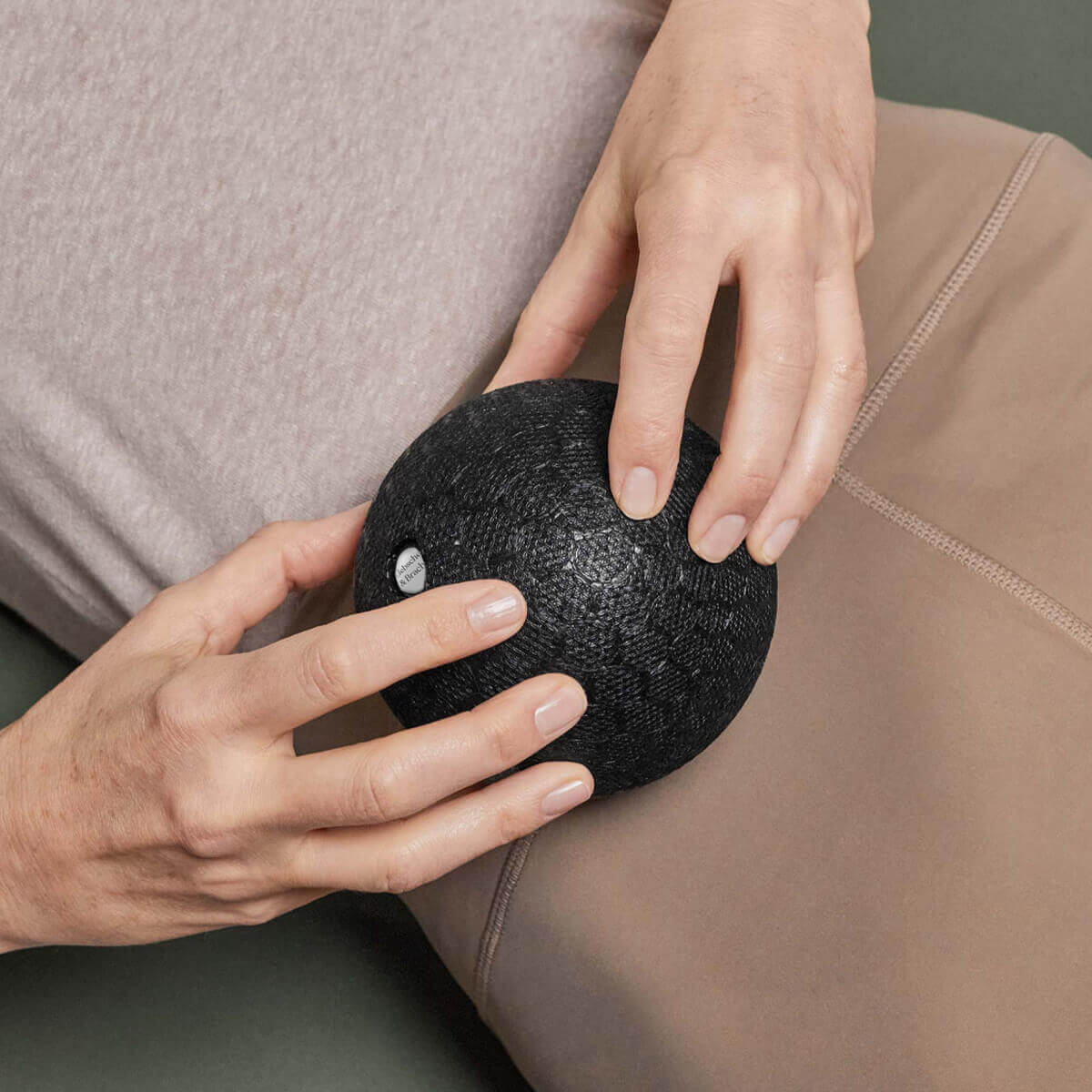 Nahaufnahme der Hände einer Person, die einen strukturierten schwarzen Massageball auf ihrem Oberschenkel hält, während sie eine beigefarbene Hose und ein hellviolettes Oberteil trägt.