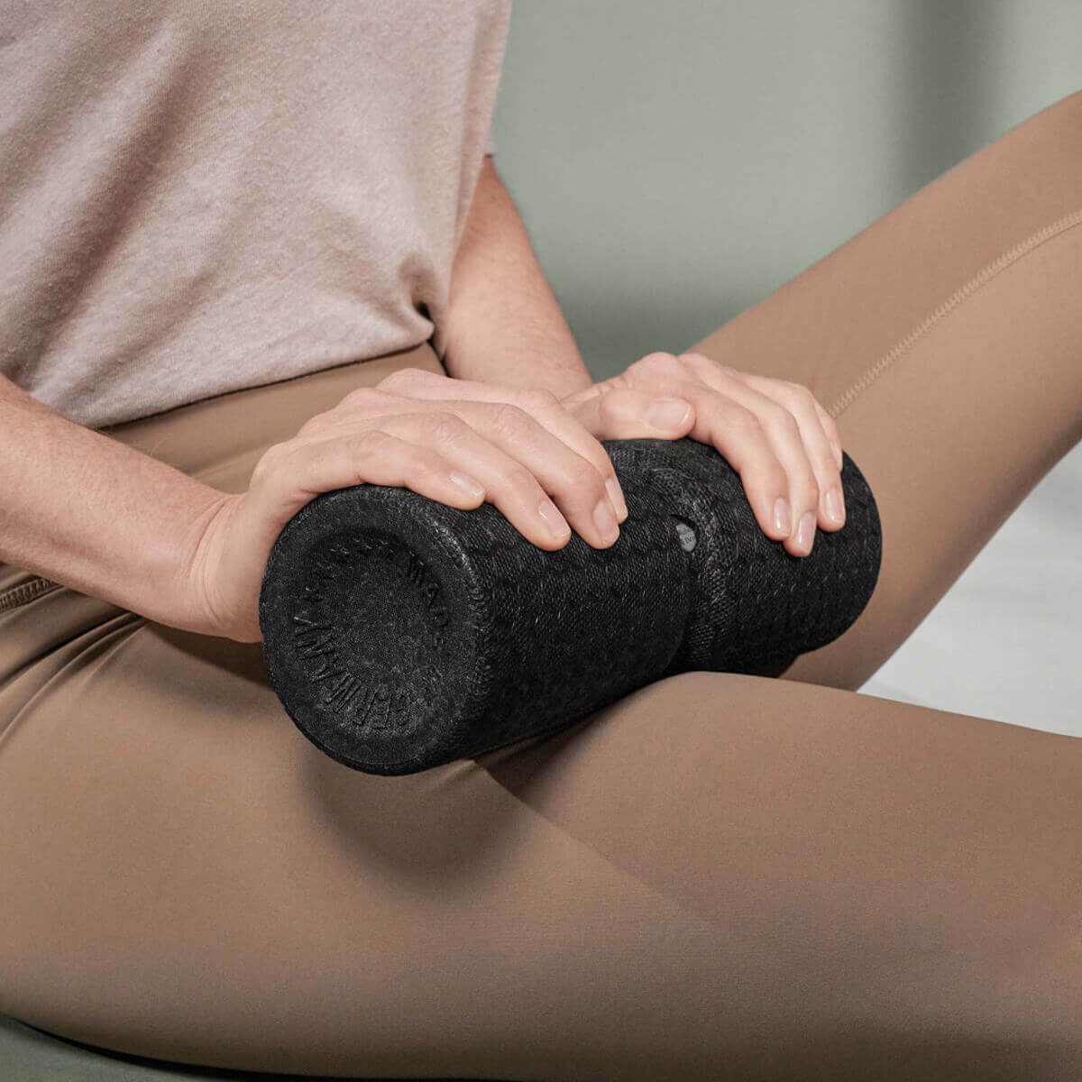 Eine Person in beigen Leggings und hellem Hemd benutzt eine schwarze Schaumstoffrolle mit Struktur auf ihrem Oberschenkel, während sie auf einer hellen Unterlage sitzt.