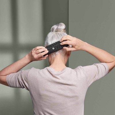 Eine Person mit grauem Haar in einem Dutt, von hinten gesehen, richtet ein schwarzes, strukturiertes Stirnband hinter ihrem Kopf, während sie ein helles Hemd vor einem gedämpften grünen Hintergrund trägt.