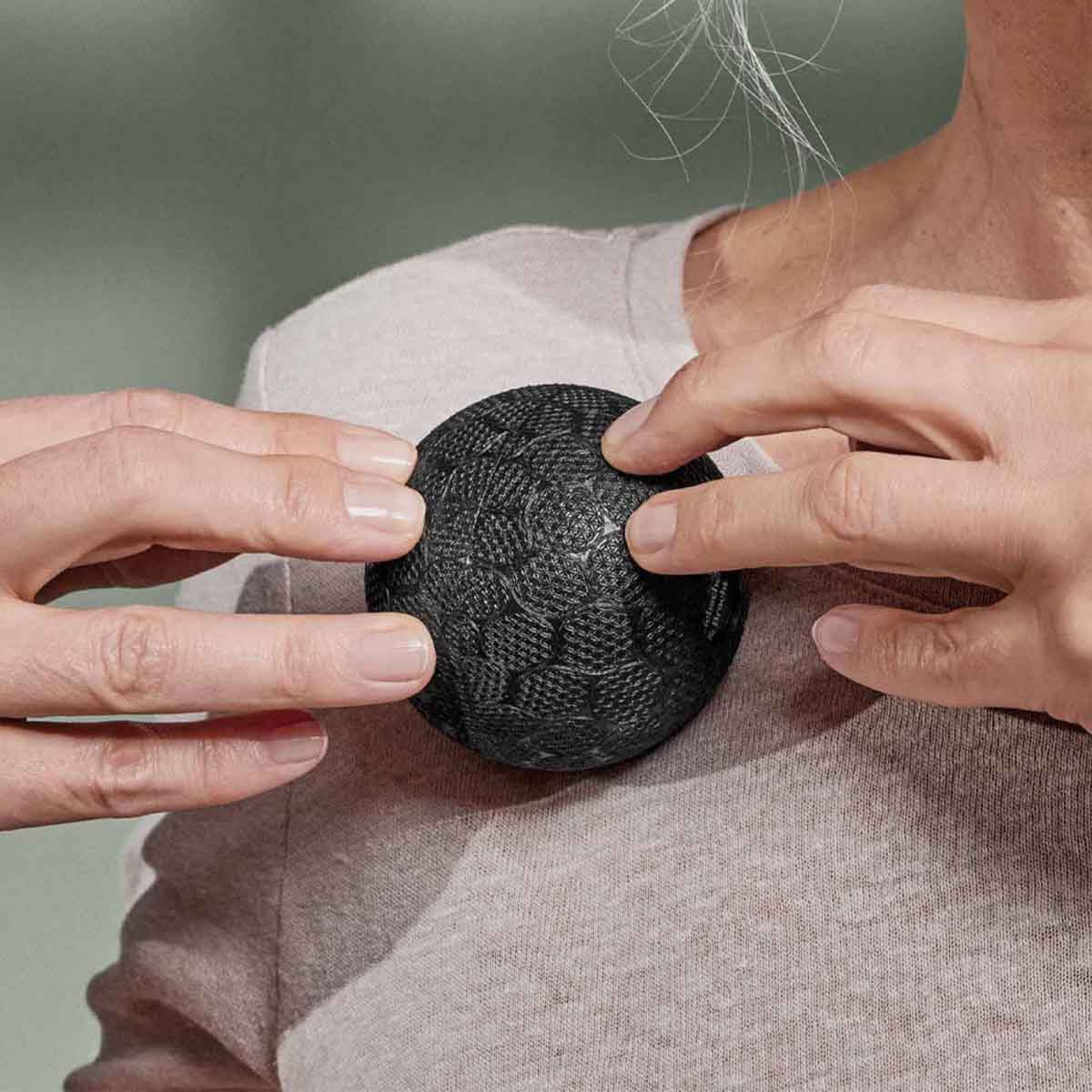 Eine Person platziert ein rundes, strukturiertes schwarzes Gerät mit einem Muster auf ihrer Brust und hält es mit beiden Händen über ein helles Hemd.