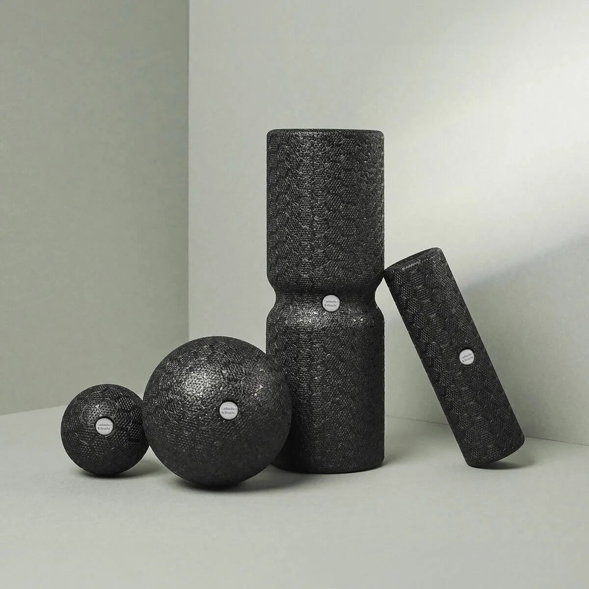 Das Liebscher & Bracht Faszien-Rollmassage-Set besteht aus vier schwarzen, texturierten Tools - zwei Bällen und zwei Zylinderrollen mit weißem Label -, ideal für Entspannung und Beweglichkeit. Präsentiert auf neutralem Hintergrund mit sanften Schatten.