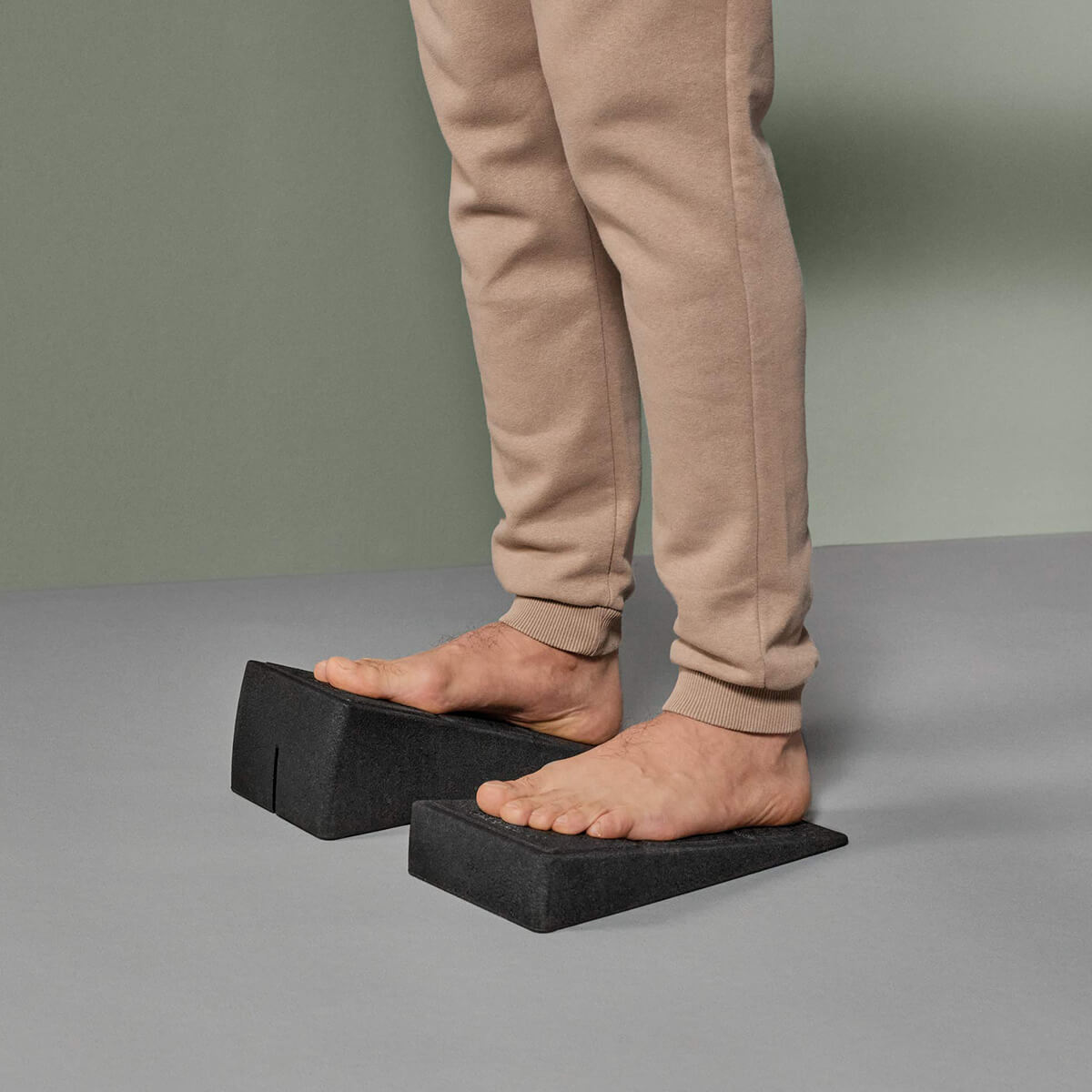 Eine Person, die eine beigefarbene Jogginghose trägt, steht barfuß auf zwei schwarzen Schaumstoffklötzen, die unter jedem Fuß auf einem grauen Boden mit gedämpftem grünem und grauem Hintergrund positioniert sind.
