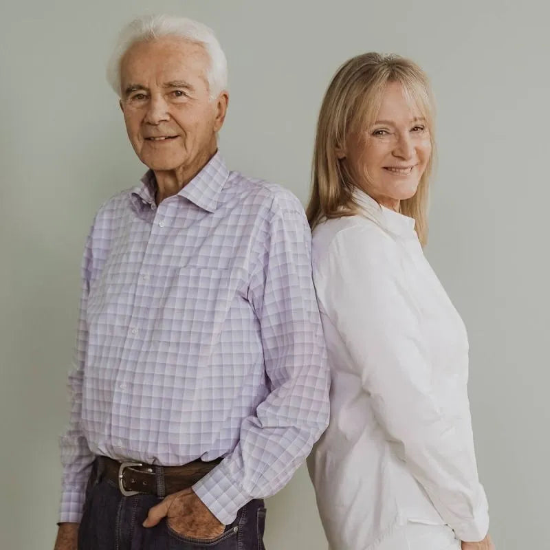 Ein älterer Mann und eine Frau stehen Rücken an Rücken und lächeln in die Kamera. Der Mann trägt ein helles kariertes Hemd und Jeans, während die Frau ein weißes Outfit trägt. Der Hintergrund ist einfarbig hell.