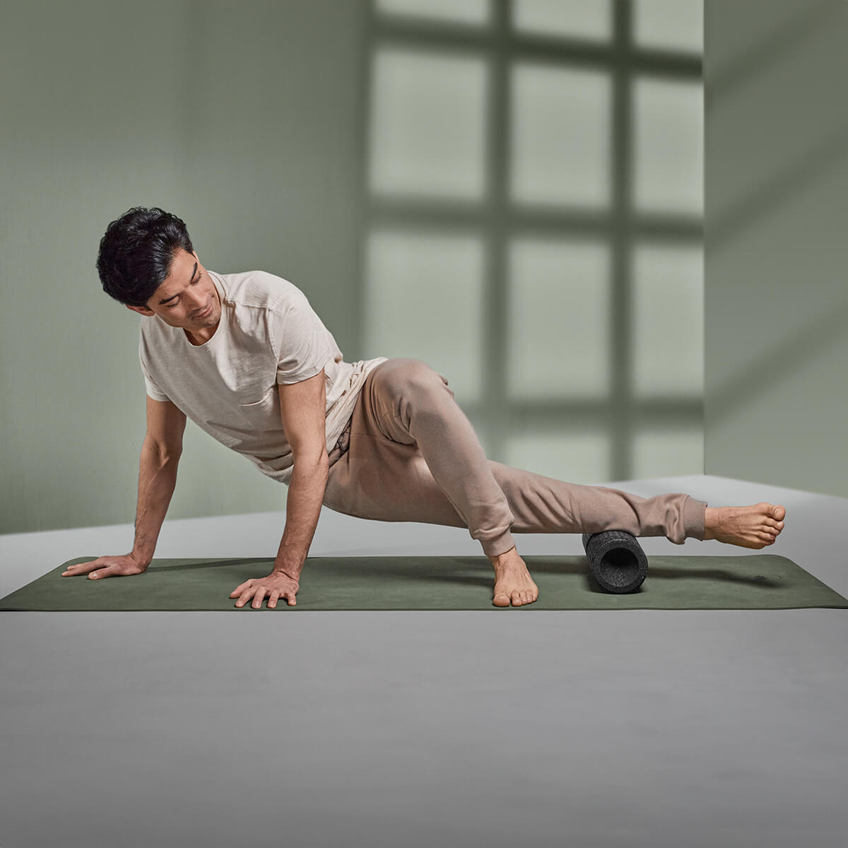Eine leicht bekleidete Person benutzt die Liebscher & Bracht MAXI-Faszienrolle mit Wirbelsäulen-Entlastung unter ihrer rechten Wade auf einer Yogamatte in einem minimalistischen Raum mit grünen Wänden und schattierten Fenstermustern, ideal für große Körperflächen.