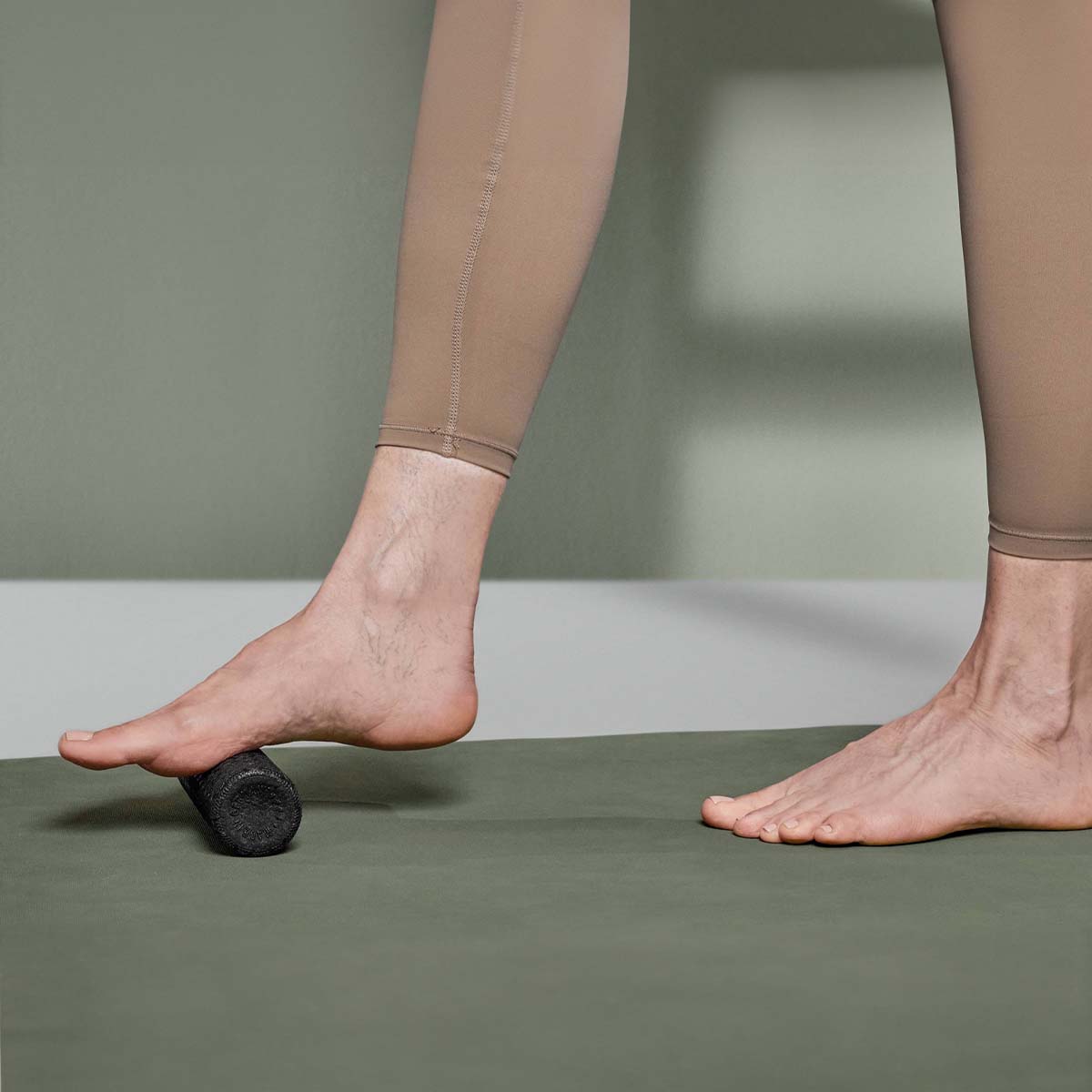 Eine Person in beigen Leggings rollt mit dem schwarzen Faszien-Rollmassage-Set von Liebscher & Bracht auf einer grünen Matte den nackten Fuß zur Entspannung und besseren Beweglichkeit durch eine wohltuende Selbstmassage.