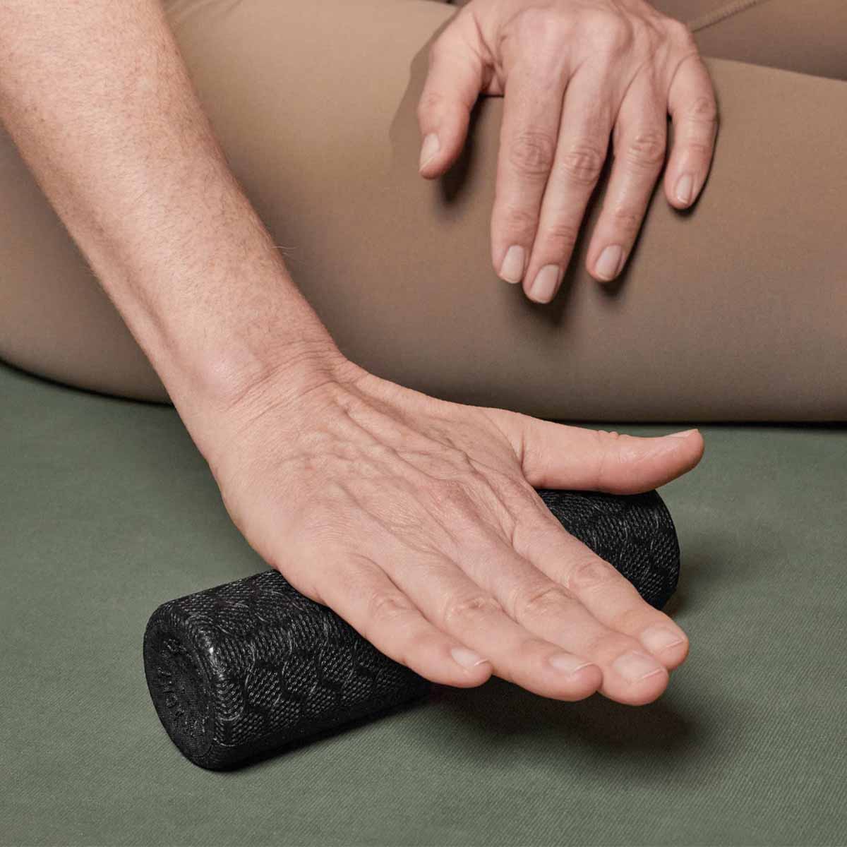 Eine Person drückt ihre Hand auf eine kleine schwarze Schaumstoffrolle, während sie im Schneidersitz auf einer grünen Matte sitzt und die andere Hand auf ihrem Bein ruht.