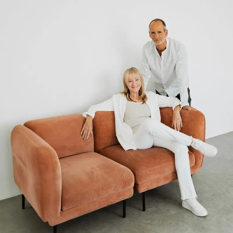Eine lächelnde Frau in weißer Kleidung sitzt auf einem hellbraunen Sofa, während ein Mann in einem weißen Hemd und einer schwarzen Hose hinter ihr steht. Beide posieren vor einer schlichten weißen Wand in einem minimalistischen Raum.