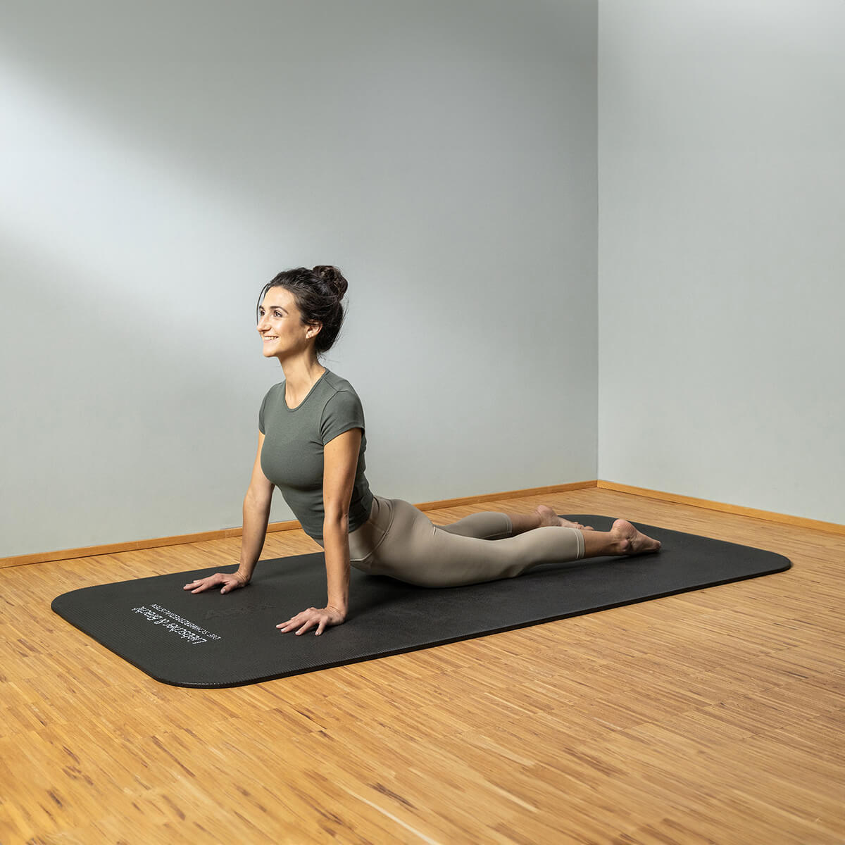 Eine Frau übt drinnen auf einer schwarzen Matte Yoga, indem sie mit gestreckten Armen, angehobener Brust und gestreckten Beinen auf einem Holzboden eine nach oben gerichtete Hundestellung einnimmt.