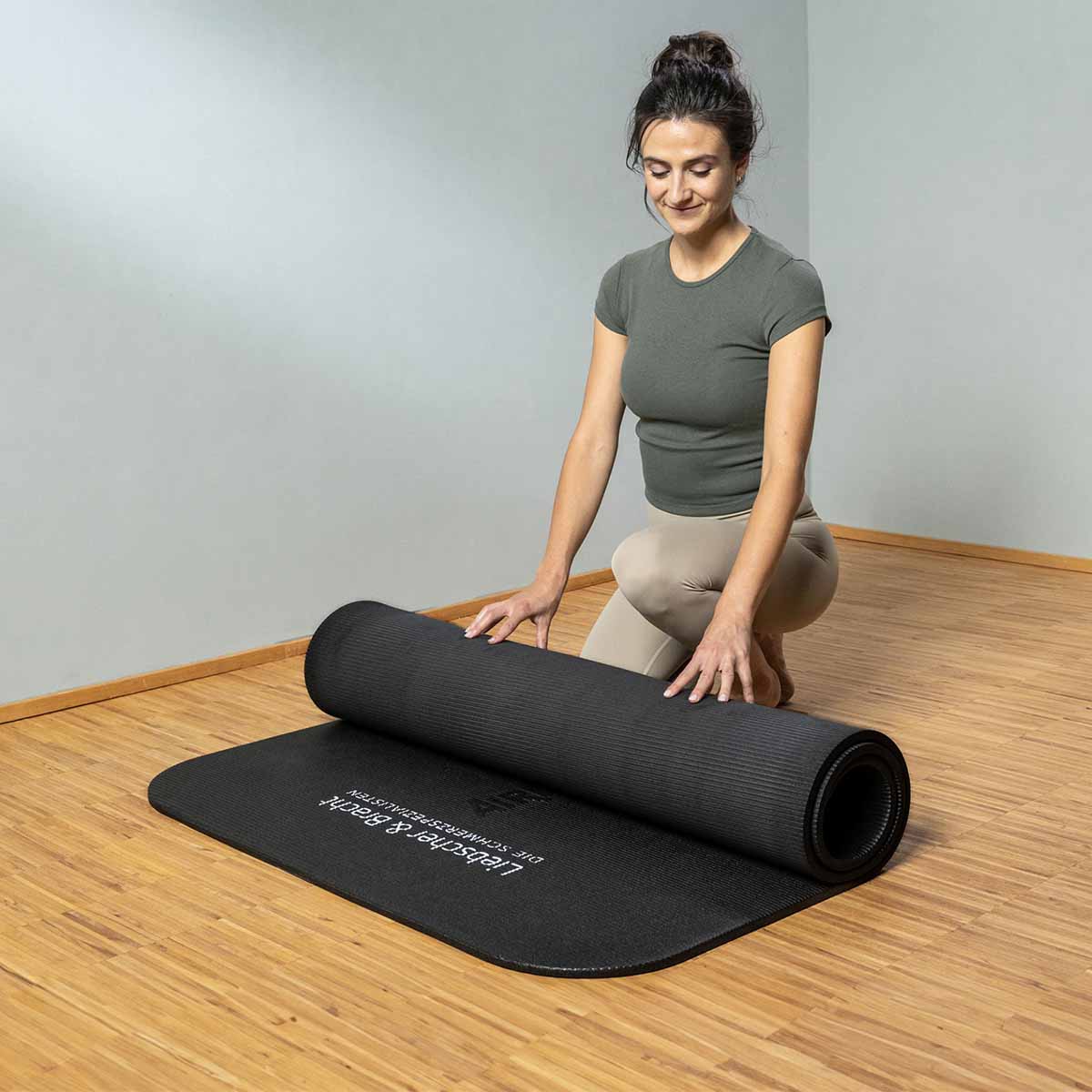 Eine Frau in einem grauen Hemd und beigen Leggings kniet auf einem Holzboden und rollt lächelnd eine schwarze Gymnastikmatte in einem hellen, minimalistischen Raum aus.