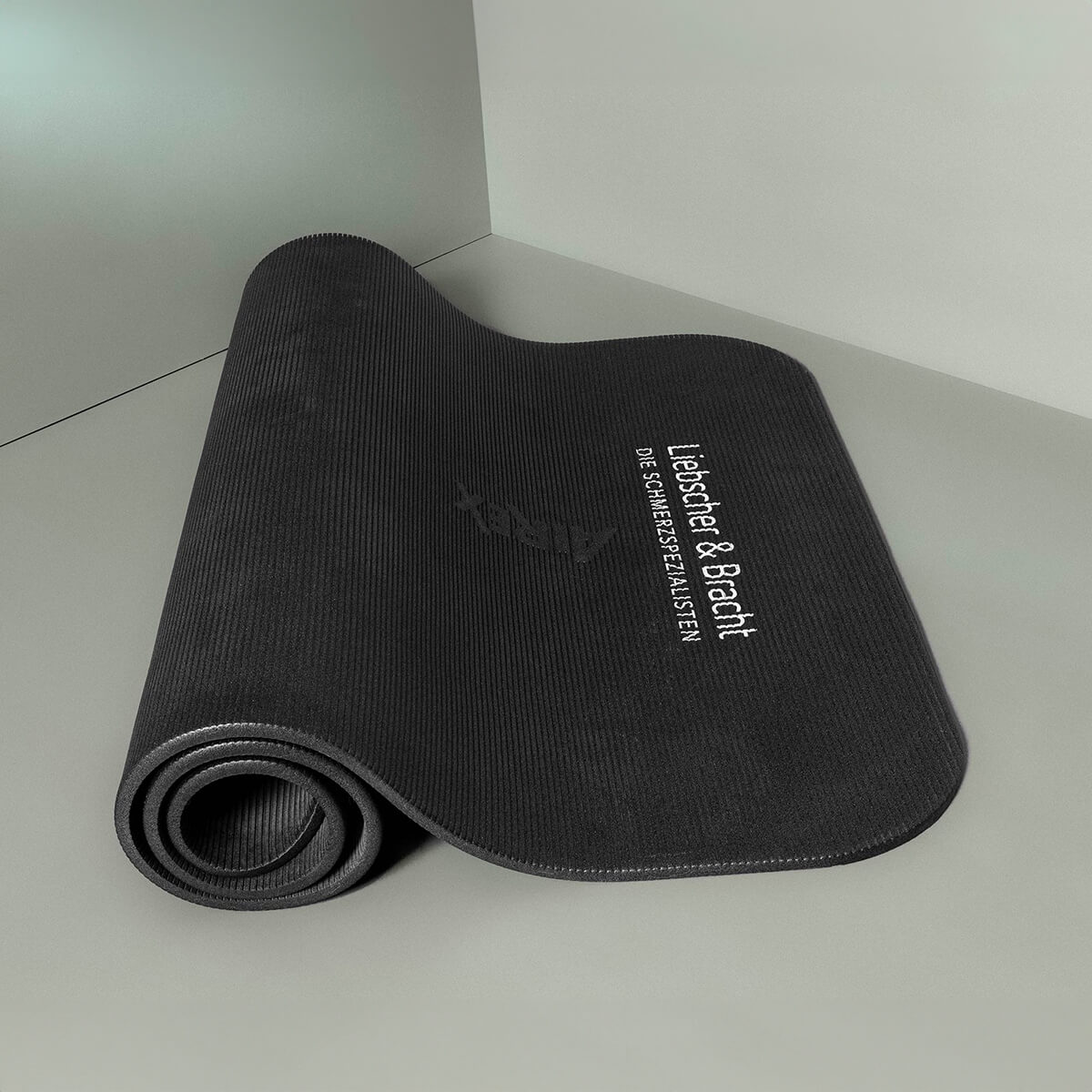 Aufgerollte schwarze Yogamatte mit strukturierter Oberfläche, die auf einem grauen Boden liegt und an einer grauen Wand lehnt. Auf der Oberfläche der Matte ist ein weißer Text zu sehen.