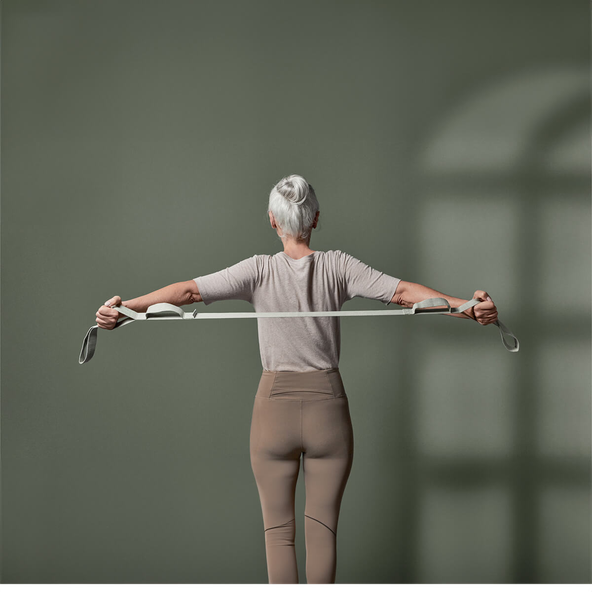 Eine Person mit grauen Haaren nutzt Der Schulterretter von Liebscher & Bracht für das Schultertraining. Sie dehnt ein Widerstandsband gegen eine grüne Wand, während sie ein helles Hemd und beigefarbene Leggings trägt und das Sonnenlicht ihren Schatten wirft.