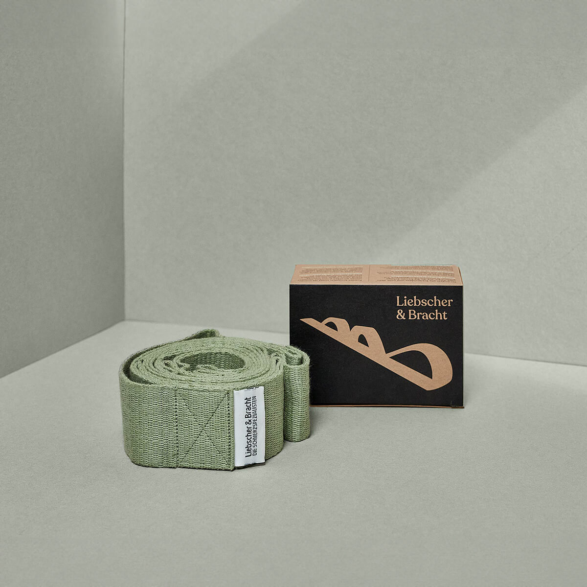 Ein sauber gerolltes hellgrünes Der Schulterretter-Band von Liebscher & Bracht mit weißem Etikett liegt neben der kleinen schwarz-braunen Schachtel auf einem minimalistischen grauen Hintergrund.