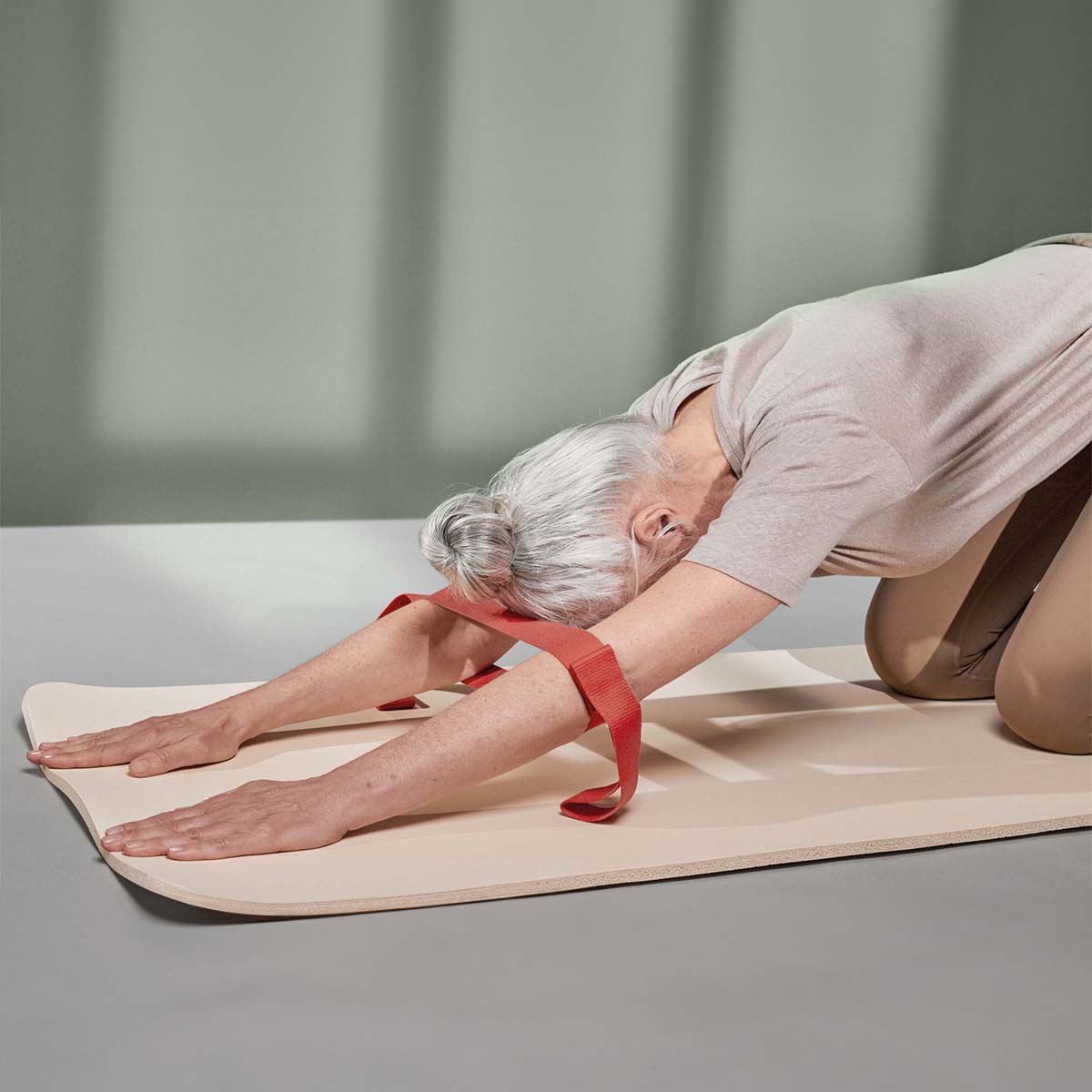 Eine ältere Frau mit grauen Haaren kniet auf einer Yogamatte und nutzt die Liebscher & Bracht Übungs-Schlaufe - ein rotes Band um ihre Arme - um Dehnpositionen zu üben und die Beweglichkeit zu verbessern.