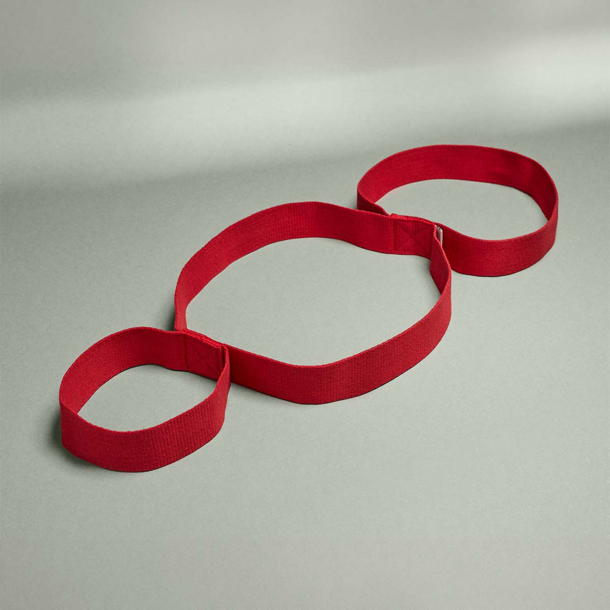 Die Liebscher & Bracht Übungs-Schlaufe, ein rotes Widerstandsband mit drei miteinander verbundenen Schlaufen für Beweglichkeit und Dehnpositionen, wird flach auf eine hellgraue Fläche gelegt.