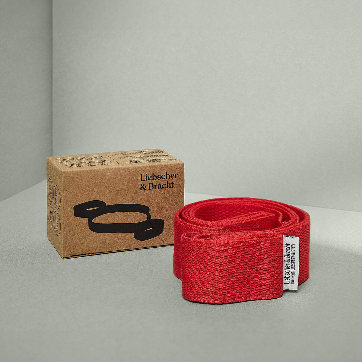 Ein rotes Übungs-Schlaufe-Stretchband von Liebscher & Bracht liegt aufgerollt neben einer braunen Schachtel mit schwarzer Grafik und Branding auf neutralem grauem Hintergrund.