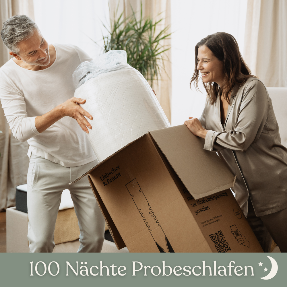 Ein lächelndes Paar packt in einem hellen Wohnzimmer eine aufgerollte Matratze aus einem großen Karton aus. Der Text am unteren Rand lautet 100 Nächte Probeschlafen mit einem Mondsichelsymbol.