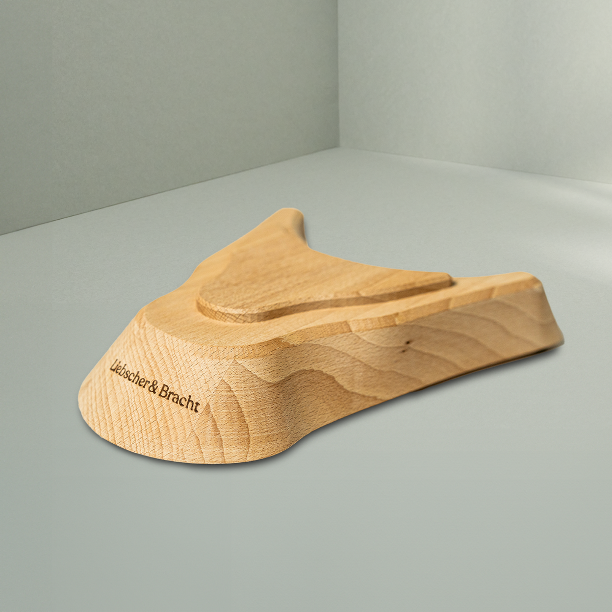 Der Liebscher & Bracht Holz-Sockel für den Nackenretter, ein Holzsockel mit sanften Rundungen und der Gravur der Marke, wird auf einer hellgrauen Fläche in einem minimalistischen Rahmen präsentiert.