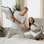 Ein lächelndes Paar im Schlafanzug genießt eine spielerische Kissenschlacht auf einem gemütlichen Bett mit Liebscher & Bracht Kissen, das für Komfort in jeder Schlafposition konzipiert ist, umgeben von Pflanzen und großen Fenstern.