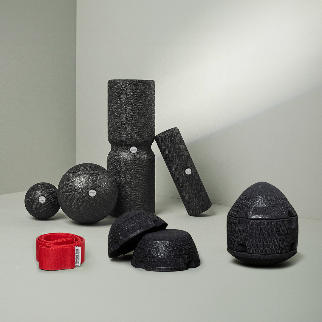 Auf einer grauen Fläche ist ein Satz schwarzer Strukturschaumrollen, Bälle und Kuppeln aus dem Dein ISG-Ischias-Paket von Liebscher & Bracht angeordnet, daneben eine rote Übungs-Schlaufe vor hellgrauem Hintergrund.
