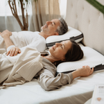 Ein Mann und eine Frau mittleren Alters liegen nebeneinander auf einem Bett in Rückenlage, zugedeckt von einer gestreiften Decke, und stützen ihre Köpfe auf Liebscher & Bracht Kissen. Sie wirken entspannt und bequem in einem sanft beleuchteten Schlafzimmer.