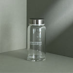 Eine Liebscher & Bracht Glasflasche mit Metalldeckel sitzt auf einer grauen Fläche, mit weißen Maßen und "Liebscher & Bracht SCHMERZSPEZIALISTEN" auf der Vorderseite - perfekt für Mixen von Pulver oder Nahrungsergänzungsmittel.