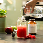 Eine Hand gießt Wasser aus einer Liebscher & Bracht Glasflasche in ein Glas mit rotem Saft auf einem Küchentisch. In der Nähe stehen grüne und rote Äpfel, rote Johannisbeeren und ein braunes Nahrungsergänzungsmittelglas mit Deckel daneben.