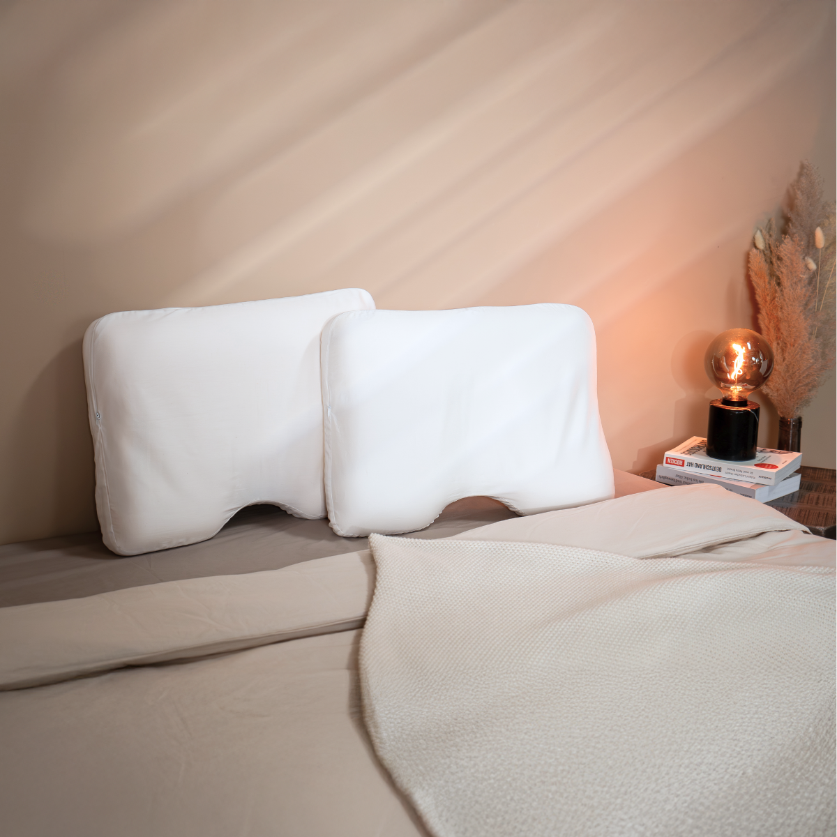Zwei weiße Liebscher & Bracht "2er-Pack: Baumwoll-Kissenbezug" liegen auf einem beigefarbenen Bett mit strukturierter weißer Decke. Auf dem Nachttisch stehen Bücher, ein Grasbouquet und eine leuchtende Kupferlampe an der beigen Wand.
