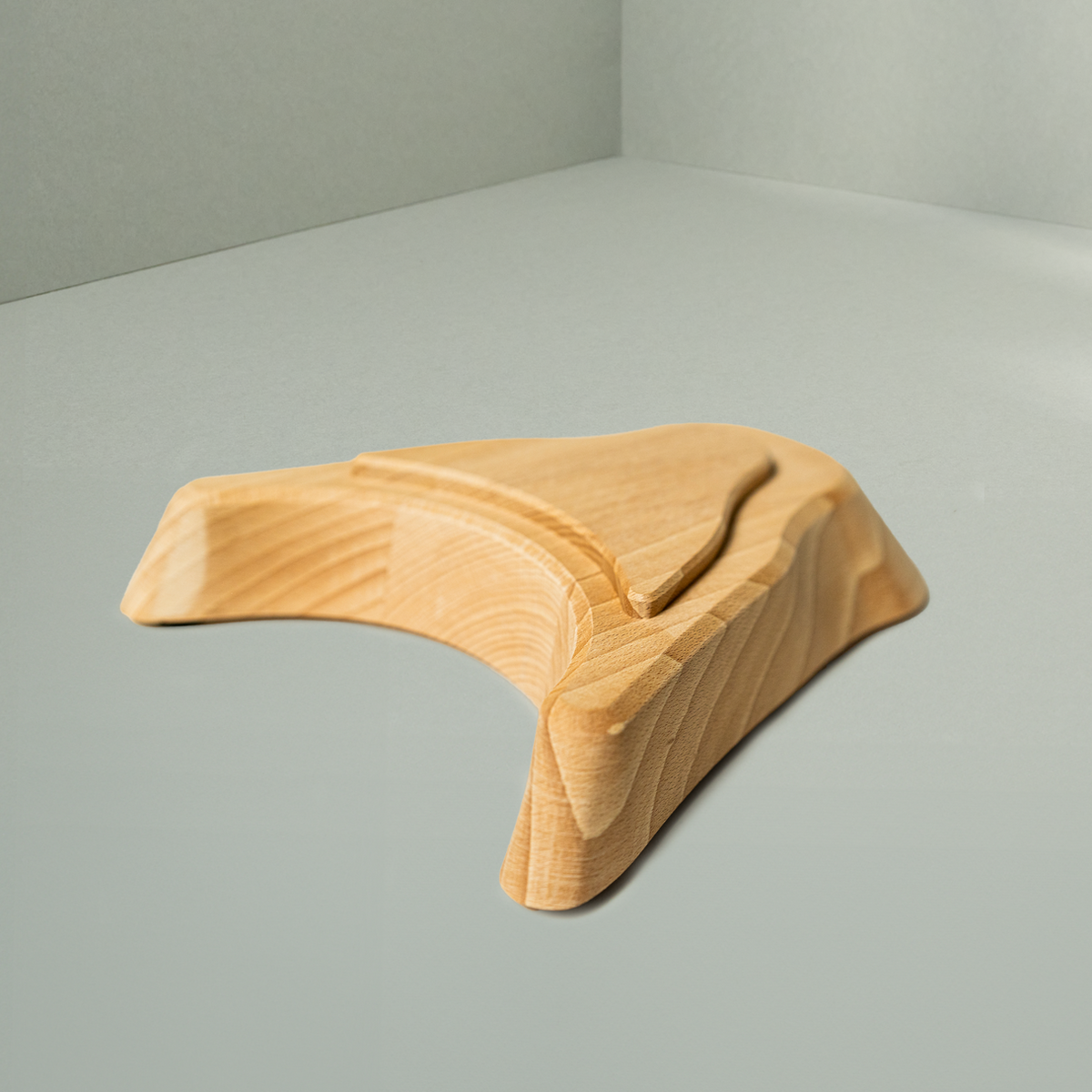 Der Liebscher & Bracht Holz-Sockel für den Nackenretter, ein glattes, helles Holzaccessoire mit geschwungenen und gewinkelten Flächen, ruht auf einer schlichten, hellgrauen Fläche in einer sanft beleuchteten Ecke.