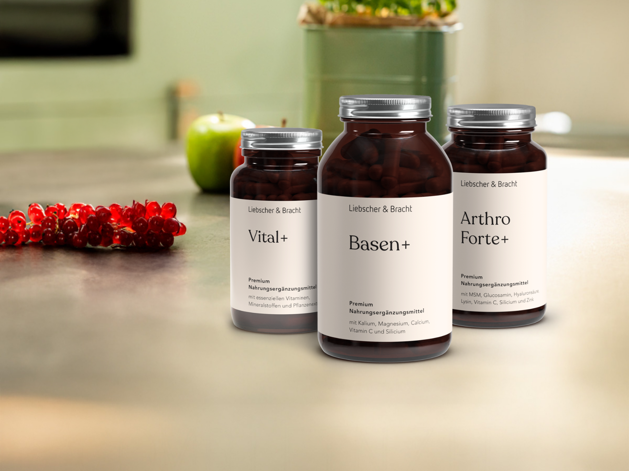 Drei Nahrungsergänzungsflaschen mit den Aufschriften Vital+, Basen+ und Arthro Forte+ stehen auf einem Küchentisch mit roten Beeren, einem grünen Apfel und einer grünen Pflanze im Hintergrund.